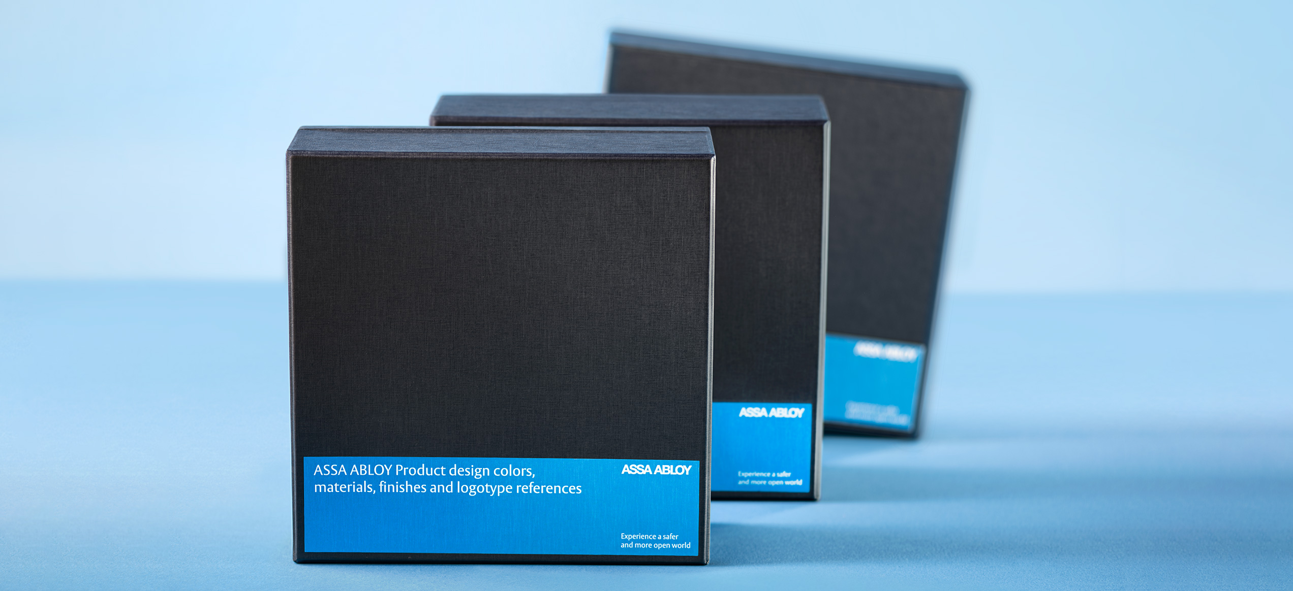 Tre scatole nere ASSA ABLOY per campionature di materiali e finiture, in fila su sfondo azzurro