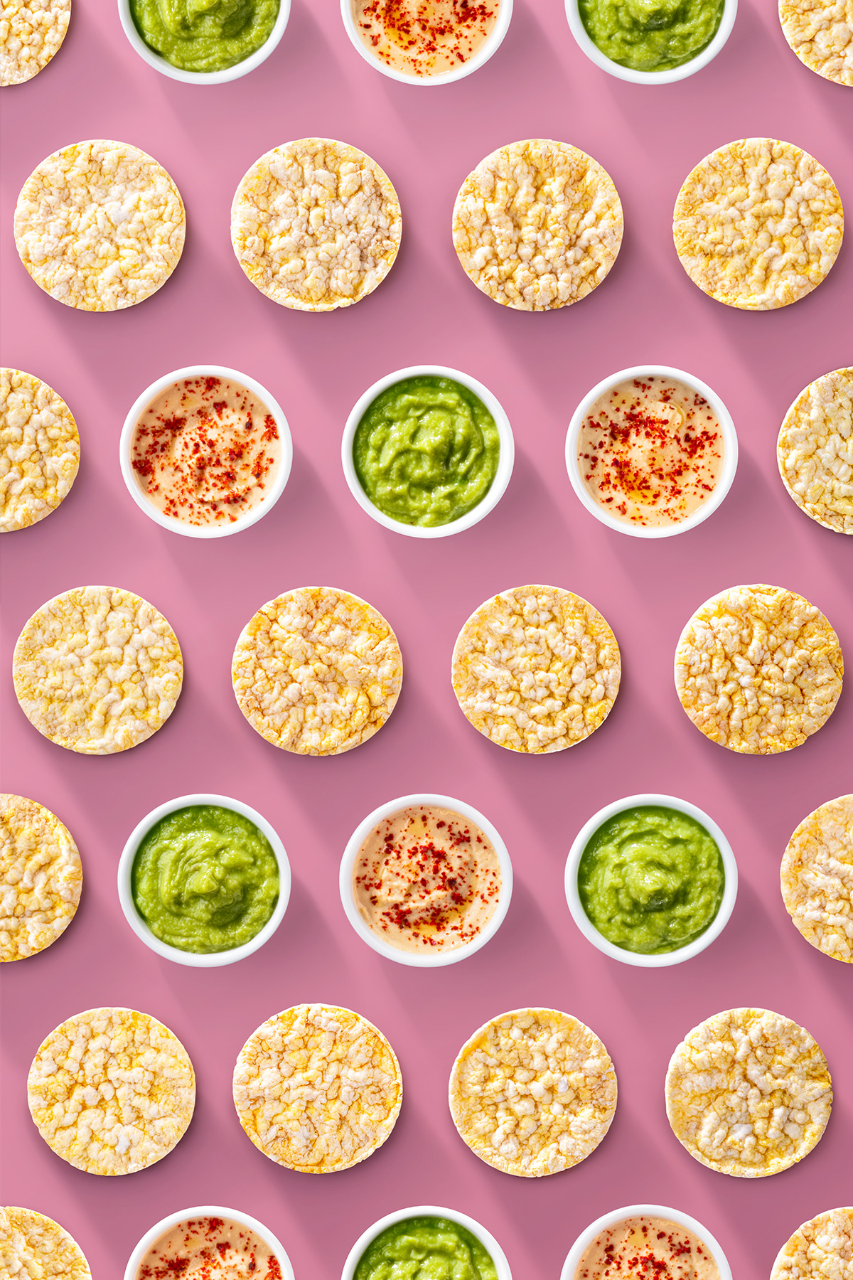 Pattern creativo con gallette Che Bon e coppette di hummus e guacamole disposte su sfondo rosa