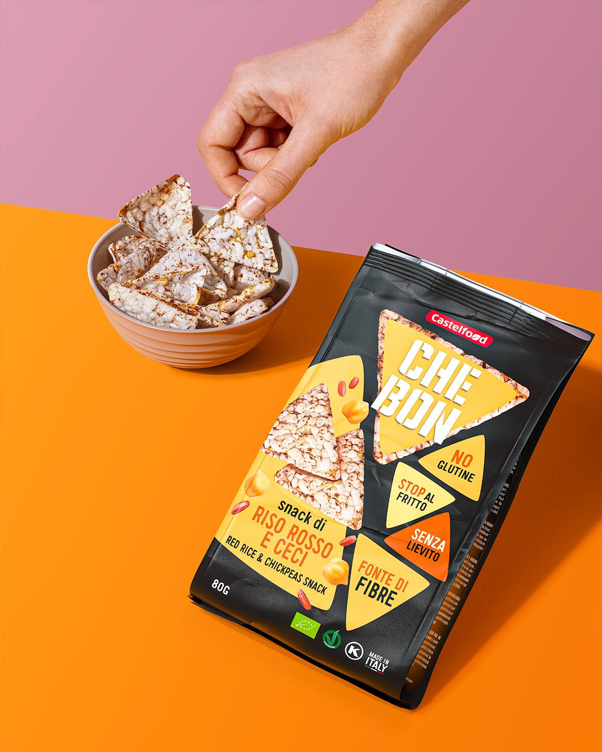Mano che prende uno snack Che Bon al riso rosso e ceci da una ciotola, con confezione a fianco su fondo arancio