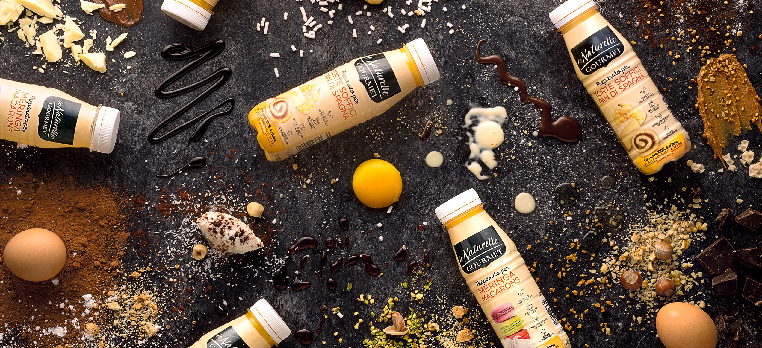 Bottiglie Le Naturelle Gourmet con ingredienti dolci su sfondo scuro: cioccolato, cocco, uova, nocciole e zuccherini