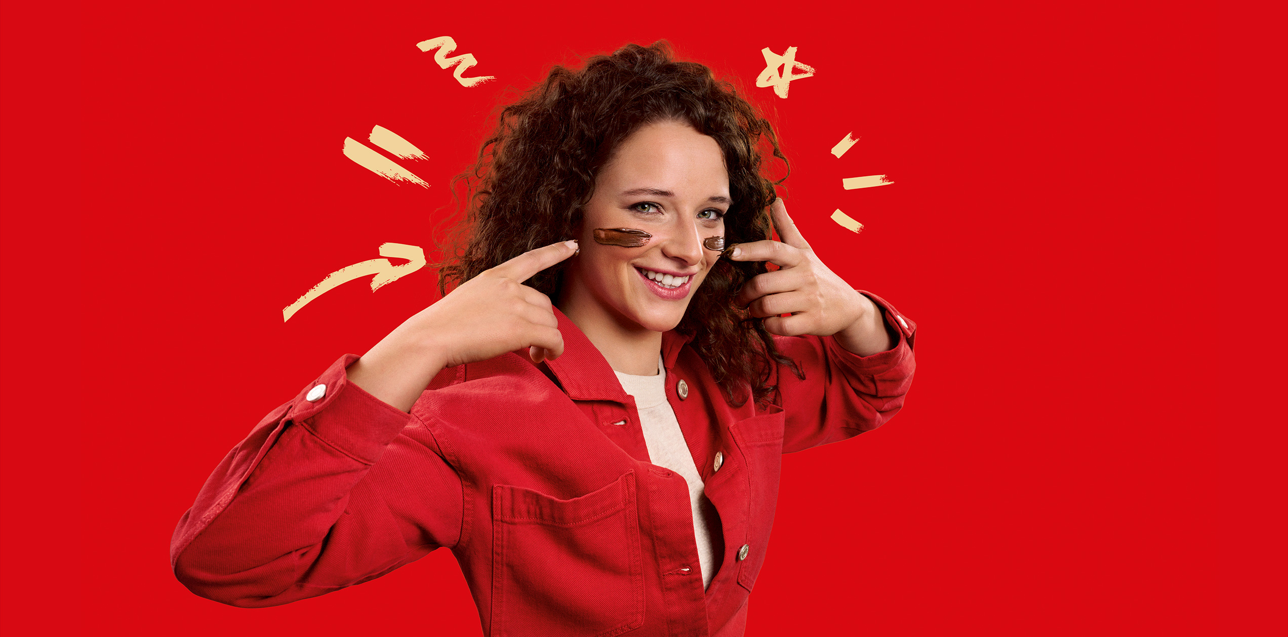 Ragazza riccia che sorride con la crema al cioccolato sulle guance, sfondo rosso e frecce disegnate che indicano il suo viso