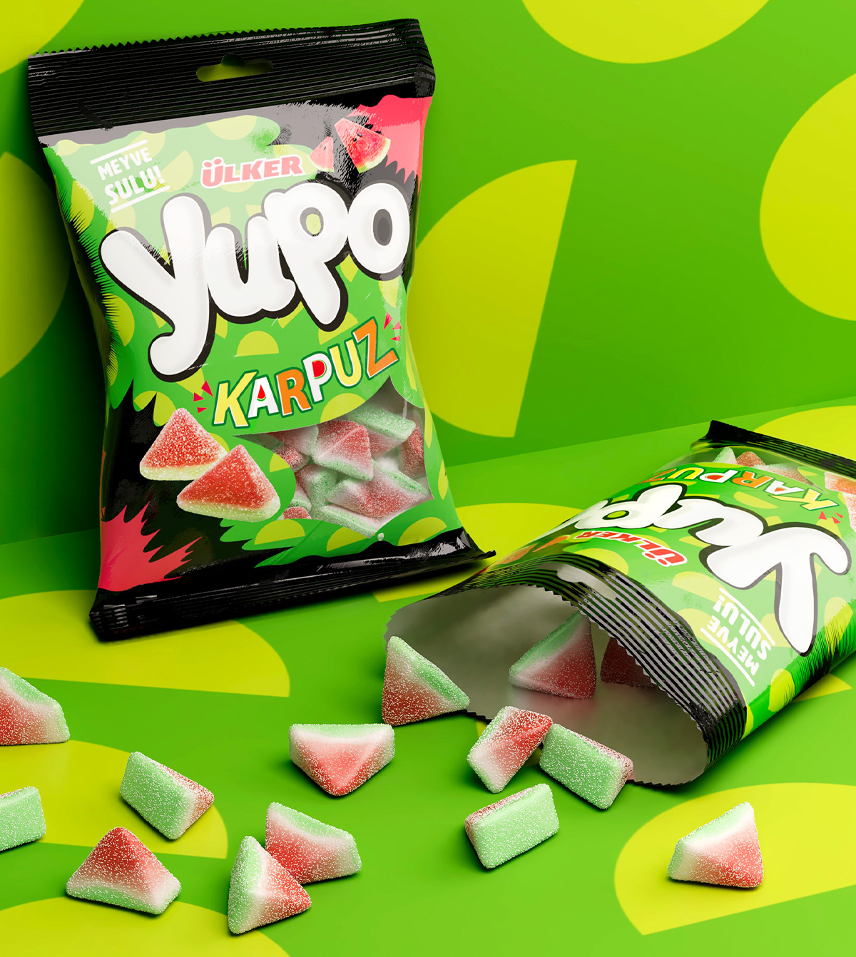 Buste di caramelle Yupo gusto anguria adagiate su uno sfondo verde acceso con pattern a mezzaluna, affiancate da caramelle triangolari sparse